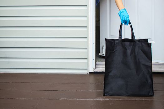 A person wearing gloves, picking up a groceries bag from a home entrance