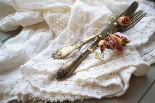 Light pastel colored tableware set: vintage silverware on napkin and delicate pink dry roses on rustic shabby wooden table