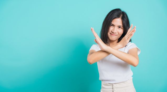 Portrait happy Asian beautiful young woman unhappy or confident standing wear white t-shirt, She holding two crossing arms say no X sign, studio shot on blue background with copy space for text