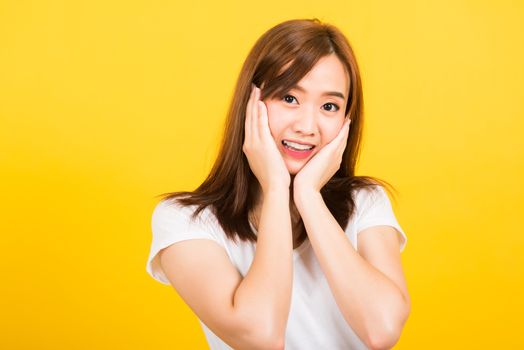 Asian happy portrait beautiful cute young woman teen stand surprised excited celebrating open mouth gesturing palms on face looking to camera isolated, studio shot yellow background with copy space