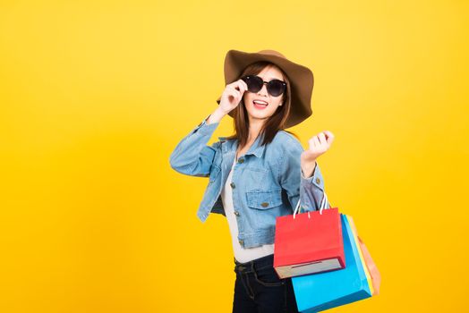 Asian happy portrait beautiful cute young woman teen smiling standing with sunglasses excited holding shopping bags multi color looking camera isolated, studio shot yellow background with copy space