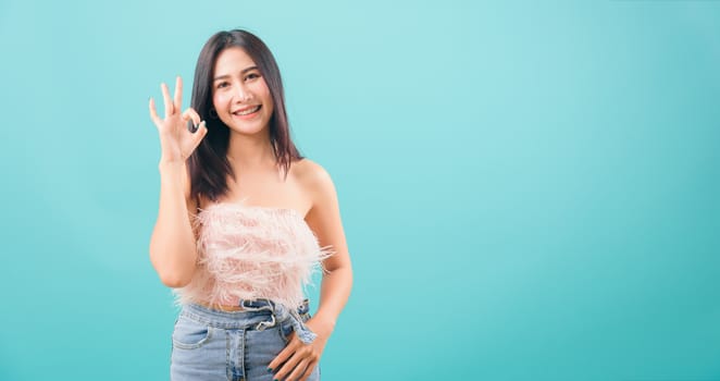 Asian happy portrait beautiful young woman standing smiling showing hand okay sign and looking to camera isolated on blue background with copy space for text