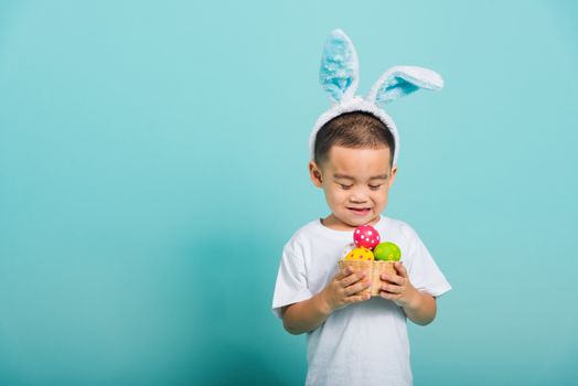 Asian cute little child boy smile beaming wearing bunny ears and a white T-shirt, standing to hold a basket with full Easter eggs on blue background with copy space