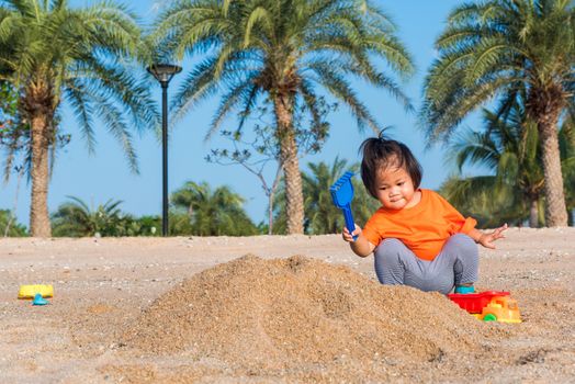 Asian Thai happy cute little cheerful daughter girl funny digging playing toy with sand at an outdoor tropical beach in summer day with copy space