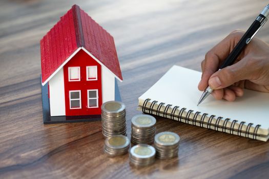 The red roof house and the pile of coins together with recording the income and expenses on the wooden table. Concept of saving money to buy a house for the future. Mortgage home and real estate.
