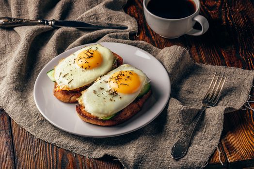 Toasts with vegetables and fried eggs on white plate and cup of coffee over grey rough cloth.