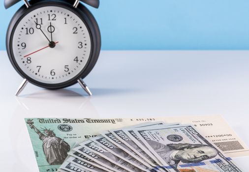 Stack of 100 dollar bills with illustrative coronavirus stimulus payment check and alarm clock to show delay in receiving money