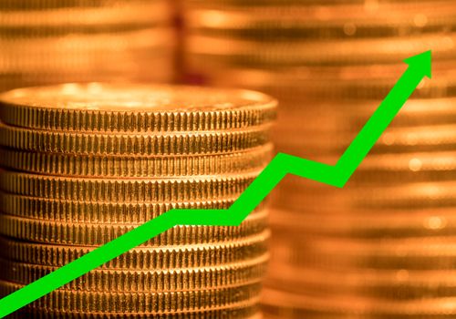 Many gold coins stacked with green arrow to illustrate the rise in price of pure gold as an investment