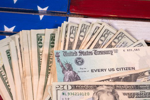 Stack of 20 dollar bills with illustrative coronavirus stimulus payment check on USA flag