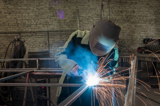 Welder welding a metal part in an industrial environment, wearing standard protection equipment. Sparks flying, fumes, industrial background