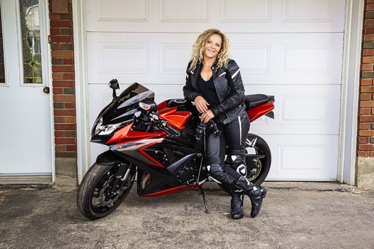 twenting someting blond woman, in full motocyclist gear, leaning on a sport motocycle, in front of a garage door