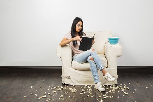 young woman sitting on a couch, reading a hardcover book and eating popcorn while making a mess
