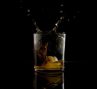 Splash in glass of whiskey and ice block falling on black background