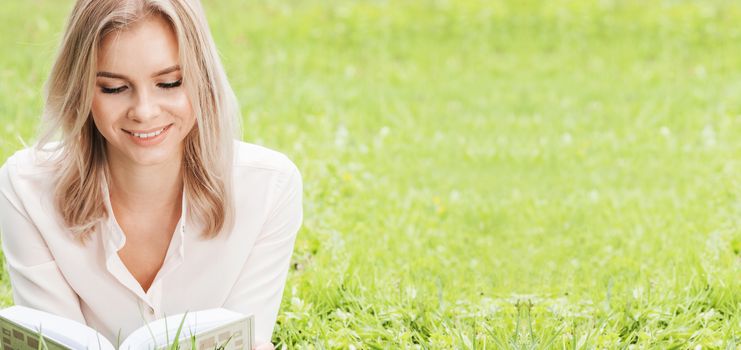 Young beautiful woman lays on green grass field and reads book, spring exam concept, copy space for text