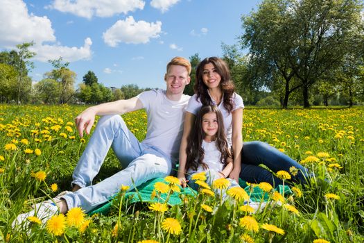 Family of three sitting on spring meadow of blooming yellow dandelion flowers being happy