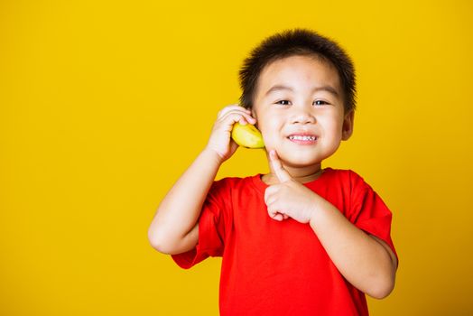 Happy portrait Asian child or kid cute little boy attractive smile wearing red t-shirt playing holds banana fruit pretending to be like a telephone, studio shot isolated on yellow background