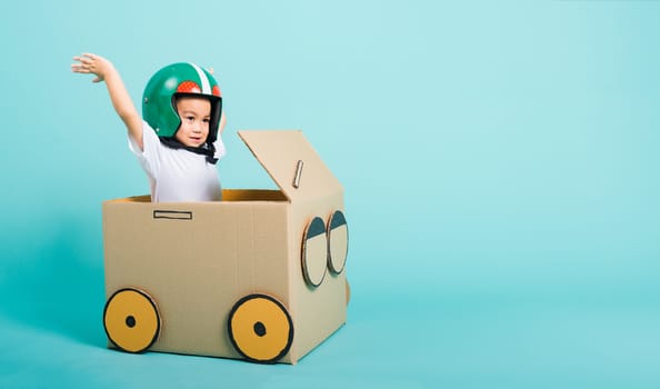 Happy Asian children boy with Helmet smile in driving play car creative by a cardboard box imagination, summer holiday travel concept, studio shot on blue background with copy space for text