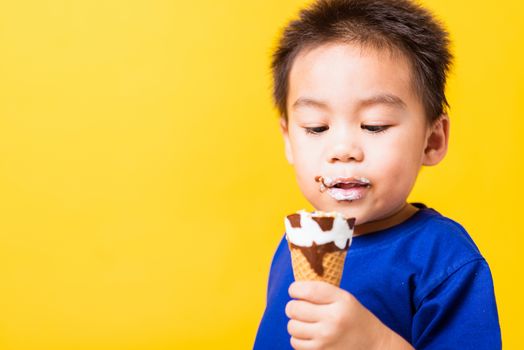 Happy portrait Asian child or kid cute little boy attractive laugh smile playing holds and eating sweet chocolate ice cream waffle cone, studio shot isolated on yellow background, summer concept