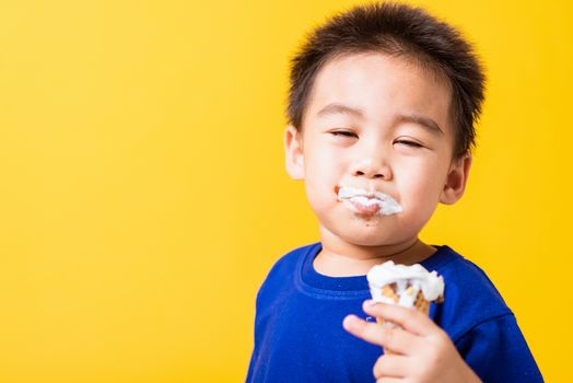 Happy portrait Asian child or kid cute little boy attractive laugh smile playing holds and eating sweet chocolate ice cream waffle cone, studio shot isolated on yellow background, summer concept