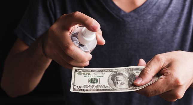 Closeup man wearing protective face mask applying sanitizer spray to 100 dollar money for cleaning, hygiene prevention COVID-19 virus or coronavirus protection concept, dark on black background