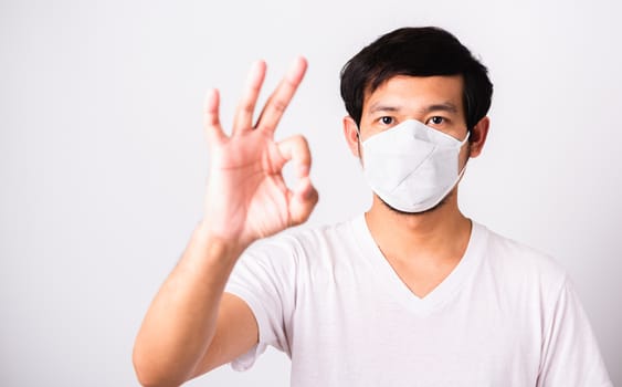 Closeup Asian handsome Man wearing surgical hygienic protective cloth face mask against coronavirus and show OK sign, studio shot isolated white background, COVID-19 medical concept