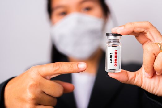 Asian woman doctor wearing surgical protective cloth face mask against coronavirus her pointing ampoule bottle vaccine focus on glass virus label, studio shot isolated white background, COVID-19