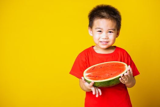 Happy portrait Asian child or kid cute little boy attractive laugh smile wearing red t-shirt playing holds cut half watermelon, studio shot isolated on yellow background