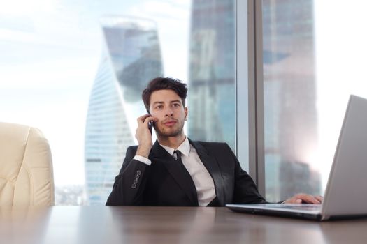 Portrait of businessman using laptop and talking on mobile phone in office