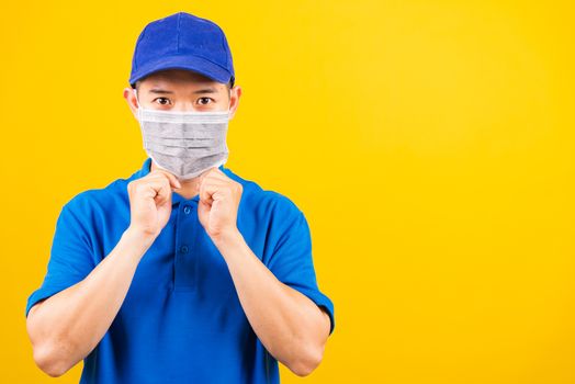 Asian young delivery worker man in blue t-shirt and cap uniform wearing face mask protective for service customer under quarantine pandemic coronavirus COVID-19, studio shot isolated yellow background