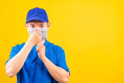 Asian young delivery worker man in blue t-shirt and cap uniform wearing face mask protective for service customer under quarantine pandemic coronavirus COVID-19, studio shot isolated yellow background