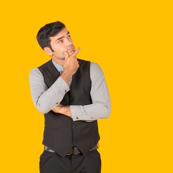 A young businessman in a gray shirt and a black vest with his hands held his chin, contemplating the project being started. Concept of investment in business. Portrait on yellow background in studio.