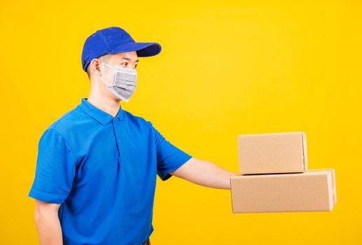 Side of Asian young delivery worker man in blue t-shirt and cap uniform wearing face mask protective giving cardboard boxes under coronavirus or COVID-19, studio shot isolated yellow background