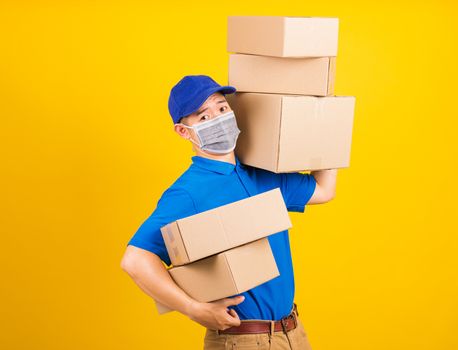 Asian young delivery worker man in blue t-shirt and cap uniform wearing face mask protective lifting stack a lot of boxes, under coronavirus or COVID-19, studio shot isolated yellow background