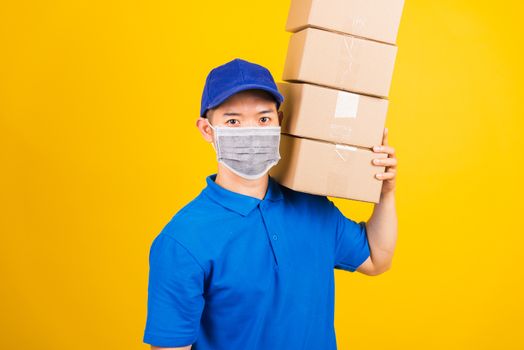 Asian young delivery worker man in blue t-shirt and cap uniform wearing face mask protective lifting stack a lot of boxes, under coronavirus or COVID-19, studio shot isolated yellow background