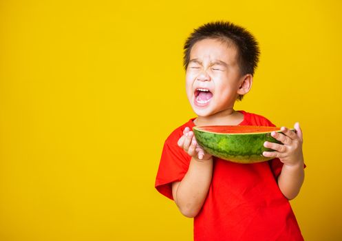 Happy portrait Asian child or kid cute little boy attractive laugh smile wearing red t-shirt playing holds cut half watermelon, studio shot isolated on yellow background