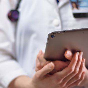 Tablet in the hands of healthcare professional up close. White coat, stethoscope, and badge visible in background. Hands of nurse practitioner or PA using technology in medicine for patients