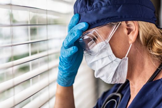 Stressed Female Doctor or Nurse On Break At Window Wearing Medical Face Mask and Goggles.