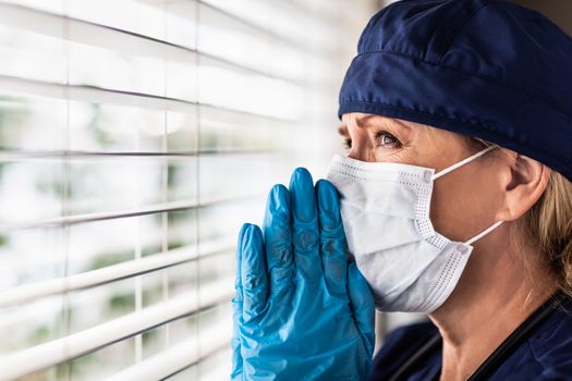 Prayerful Stressed Female Doctor or Nurse On Break At Window Wearing Medical Face Mask.