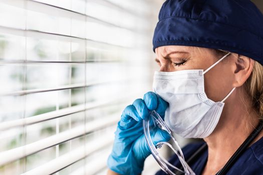 Stressed Female Doctor or Nurse On Break At Window Wearing Medical Face Mask and Goggles.