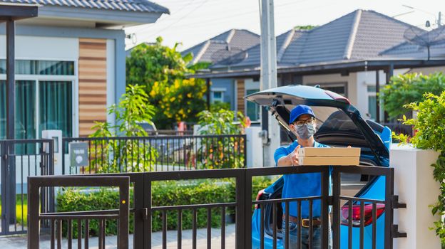 Asian Young delivery man courier online with box in blue cap and t-shirt uniform he protective face mask, service customer in front of the house under curfew quarantine pandemic coronavirus COVID-19