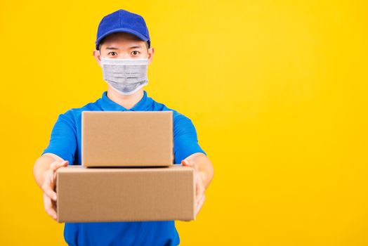 Front of Asian young delivery worker man in blue t-shirt and cap uniform wearing face mask protective giving cardboard boxes under coronavirus or COVID-19, studio shot isolated yellow background