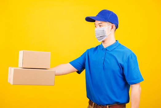 Side of Asian young delivery worker man in blue t-shirt and cap uniform wearing face mask protective giving cardboard boxes under coronavirus or COVID-19, studio shot isolated yellow background