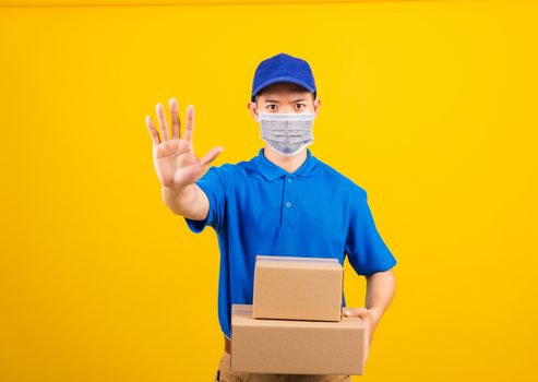 Asian young delivery worker man in blue t-shirt and cap uniform wearing face mask protective hold boxes he raise hand to stop sign under coronavirus or COVID-19, studio shot isolated yellow background