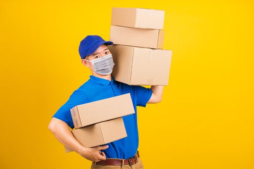 Asian young delivery worker man in blue t-shirt and cap uniform wearing face mask protective lifting stack a lot of boxes, under coronavirus or COVID-19, studio shot isolated yellow background