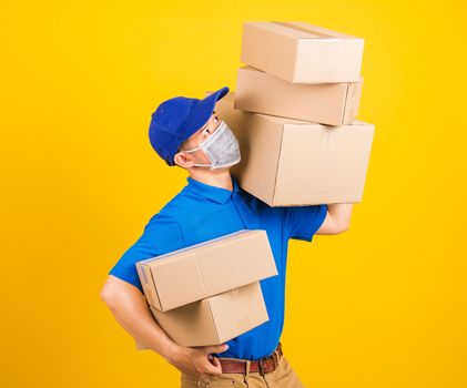 Asian young delivery worker man in blue t-shirt and cap uniform wearing face mask protective lifting stack a lot of boxes, under coronavirus or COVID-19, studio shot isolated yellow background