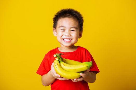 Happy portrait Asian child or kid cute little boy attractive smile wearing red t-shirt playing holds bananas comb fruit, studio shot isolated on yellow background