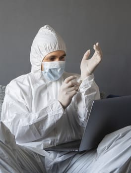 Man in protective suit, medical mask and rubber gloves sits at home and works with laptop at the table during quarantine. Designer, artist, architect, businessman at remote work in a pandemic covid