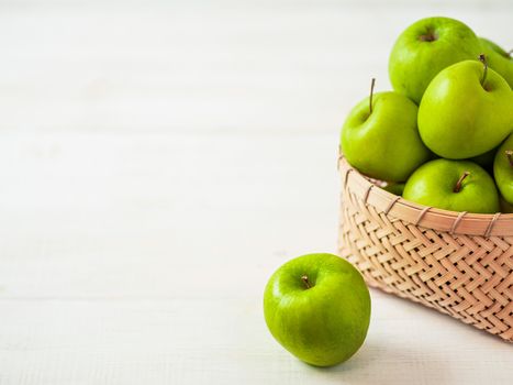 Apples harvest concept. Braided Bamboo Basket with green apples on white wooden background with copy space for text or design.