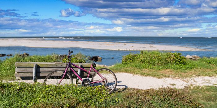 An old purple bike leaning against an empty bench on a beach by a blue sea