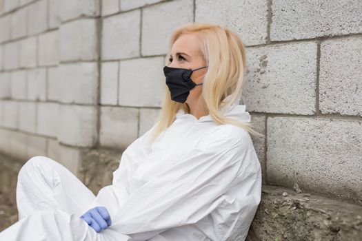 Woman in black mask, white protective suit and gloves sitting against wall